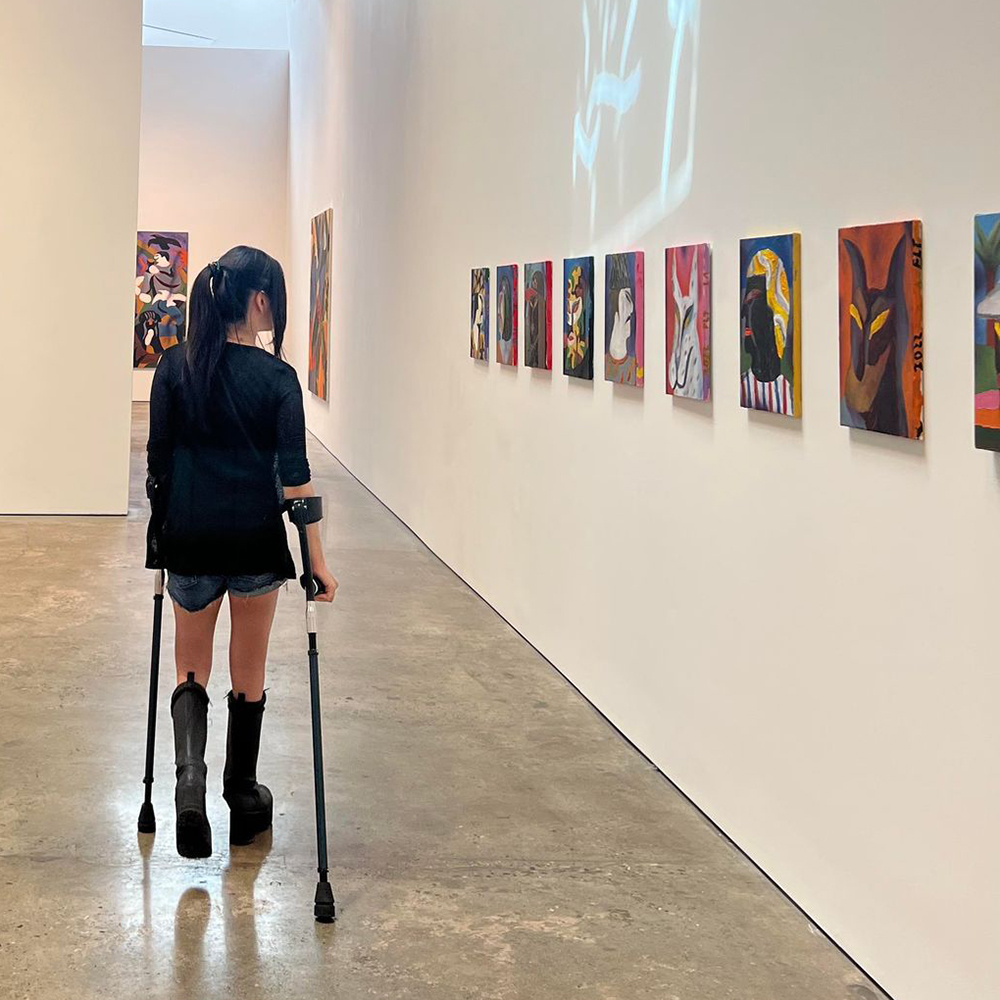 Me walking through an art gallery looking at the paintings on the wall. I'm looking and walking away from the camera and I have my straight black hair up in a ponytail. I'm wearing a red tanktop, black cardigan, denim shorts, and black combat boots, and using color-shifting forearm crutches.